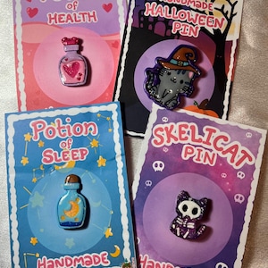 May include: Four handmade enamel pins on cards. The pins include a potion of health, a Halloween cat, a potion of sleep, and a skelcat. The cards have a pink, purple, blue, and purple background with the words "Potion of Health", "Handmade Halloween Pin", "Potion of Sleep", and "Skelicat Pin".