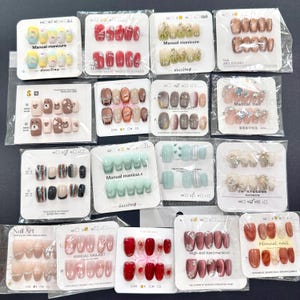 May include: A collection of artificial nails in various designs and colors, including French tips, glitter, and patterned styles. The sets are packaged on individual cards, with some labeled "Manual manicure" and "Nail Art".