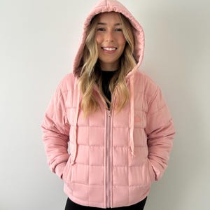 Könnte beinhalten: Hellrosa gesteppte Jacke mit Kapuze und Frontreißverschluss. Die Jacke hat ein quadratisches Muster und Kordelzüge an der Kapuze. Das Model trägt eine schwarze Hose.