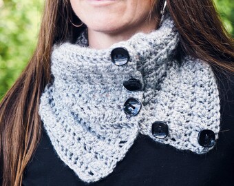May include: A gray crocheted cowl with a pointed collar and four black buttons. The cowl is worn by a person with long brown hair.