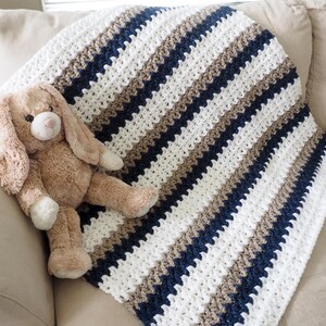 May include: A white, brown, and navy blue striped crocheted baby blanket with a plush brown bunny toy on top.