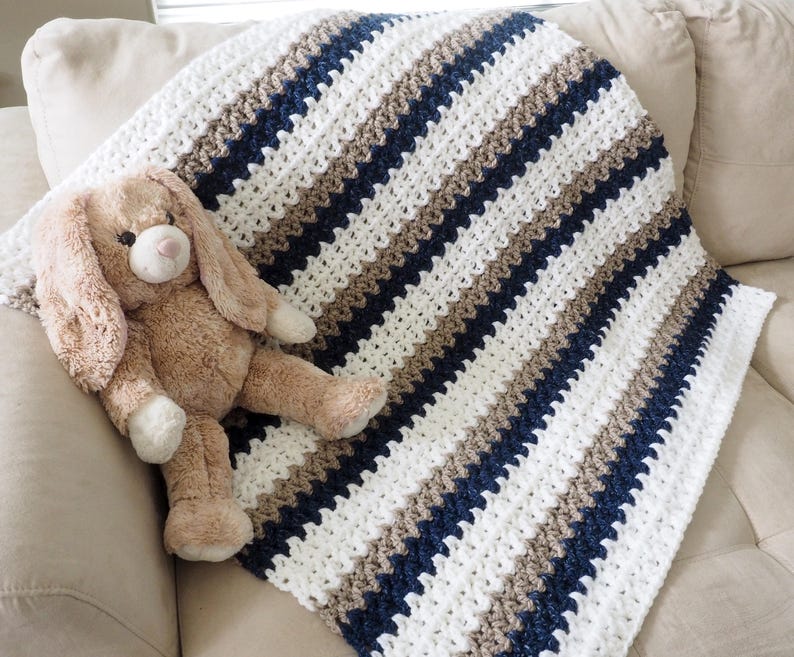 May include: A white, brown, and navy blue striped crocheted baby blanket with a plush brown bunny toy on top.