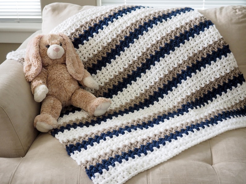 May include: A white, brown, and navy blue striped crocheted blanket with a plush brown bunny toy on top.