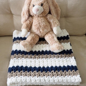 May include: A white, brown, and navy blue striped crocheted baby blanket with a plush brown bunny toy sitting on top.