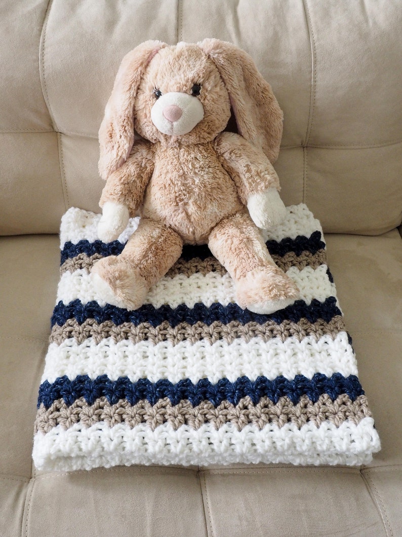 May include: A white, brown, and navy blue striped crocheted baby blanket with a plush brown bunny toy sitting on top.