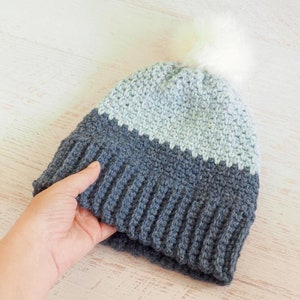 May include: A gray and light blue crocheted beanie with a white pom pom on top.
