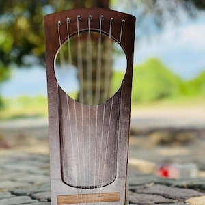 May include: A wooden lyre with a dark brown finish. The instrument has a large circular sound hole and multiple strings. The lyre is set against a blurred natural background, showcasing its elegant design. The lyre is a traditional stringed instrument.