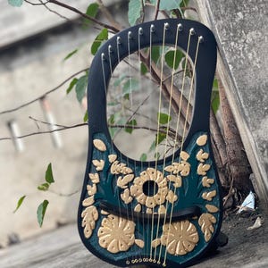 Può includere: Una lira nera con disegni floreali dorati su sfondo verde acqua. Lo strumento ha corde dorate ed è appoggiato a un muro di cemento con rami di alberi sullo sfondo. La lira è uno strumento musicale a corde.