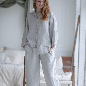 以下が含まれることがあります： 襟付きの長袖ボタンダウンシャツと、お揃いのワイドレッグパンツで構成されたライトグレーのツーピースラウンジウェアセット。シャツはフロントボタン留めで、パンツにはサイドポケットが付いており、どちらも質感のある生地で作られています。