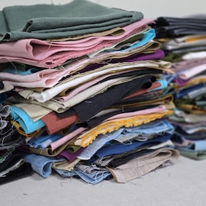 May include: A stack of fabric scraps in various colors, including green, pink, blue, and black. The fabric pieces are layered and appear to be of different textures and weights, suitable for quilting or other craft projects.