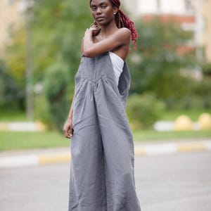 Könnte beinhalten: Eine Person trägt einen grauen Overall mit weiten Beinen und einem weißen Oberteil. Der Overall hat eine lockere Passform und ist aus einem weichen Stoff gefertigt. Sie trägt Sandalen. Der Hintergrund ist verschwommen, mit Bäumen und einem Gebäude.