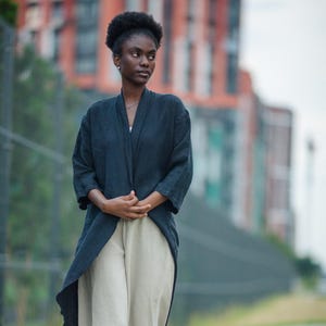 May include: A woman wearing a dark navy blue kimono-style jacket over light beige linen pants. The jacket has a long, asymmetrical hem. The pants are wide-legged. Black clogs with decorative elements complete the outfit. The background features a blurred urban setting.