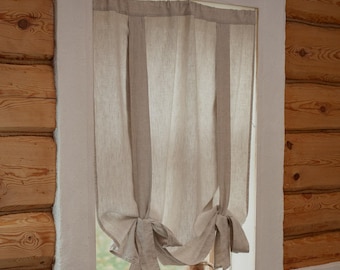 May include: A light beige linen curtain hanging in a window, featuring two vertical fabric ties that are knotted at the bottom to create a gathered, draped effect. The fabric has a natural, textured weave and hangs against a white window frame.