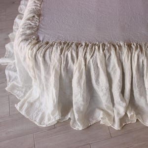 May include: A close-up of a bed with a white ruffled bed skirt. The bed skirt has a gathered design and hangs down to the floor. The bedspread is a light color and has a textured appearance.