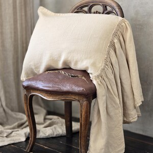 Pode incluir: Uma almofada de linho bege e tecido a condizer drapeados sobre uma cadeira de madeira vintage com um assento de couro castanho desgastado. A cadeira tem pernas curvas e um encosto decorativo, num fundo neutro.