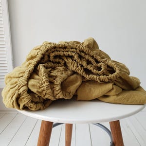May include: Mustard-colored fitted sheet with elastic edges, neatly folded on a white round table. The sheet is made of a soft, textured fabric, likely linen or cotton, and is a solid color. The sheet is a neutral tone.