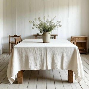 May include: A bright, airy dining room with white walls and a light wood floor. A large wooden table is covered with a white linen tablecloth. A vase of flowers sits in the center. A metal pendant light hangs above the table.
