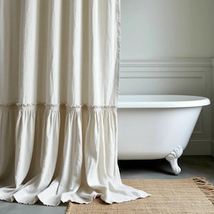 Peut inclure: Un rideau de douche crème avec un bord à volants est suspendu devant une baignoire blanche à pieds de lion. Le rideau est en tissu texturé. Un tapis en jute tressé est posé sur le sol devant la baignoire.