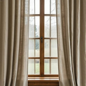 May include: Beige linen curtains frame a wooden window with multiple panes. The window looks out onto a blurred outdoor scene with trees and grass. The curtains are draped and create soft folds.