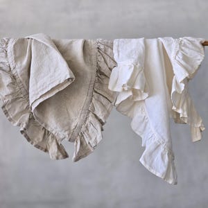 May include: Two linen textiles with ruffled edges are draped over a wooden rod. One is a light beige color, and the other is white. The ruffles add a decorative element to the otherwise simple design. The background is a solid, neutral gray.