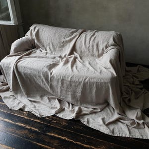 May include: A light beige linen sofa throw blanket draped over a couch. The throw blanket is made of natural linen fabric and has a relaxed, casual look.