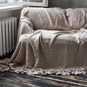 Peut inclure: Une housse de canap&eacute; en lin beige dot&eacute;e d'un bord &agrave; volants le long de l'ourlet inf&eacute;rieur. Le tissu pr&eacute;sente une texture tiss&eacute;e visible et se drape librement sur le meuble, descendant jusqu'au sol avec des plis souples et un aspect naturel.