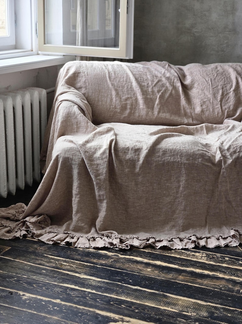 Peut inclure: Une housse de canap&eacute; en lin beige dot&eacute;e d'un bord &agrave; volants le long de l'ourlet inf&eacute;rieur. Le tissu pr&eacute;sente une texture tiss&eacute;e visible et se drape librement sur le meuble, descendant jusqu'au sol avec des plis souples et un aspect naturel.