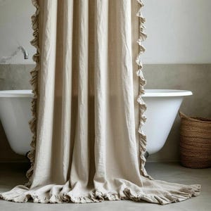 Könnte beinhalten: Beigefarbener Duschvorhang aus Leinen mit Rüschenkanten. Der Vorhang hängt vor einer weißen Badewanne, rechts ist ein Weidenkorb zu sehen. Der Stoff des Vorhangs fällt auf den Boden.