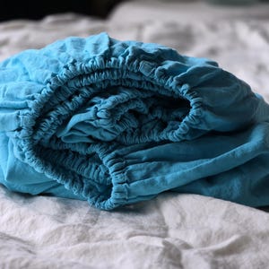 Peut inclure: Un drap-housse turquoise plié, avec des bords élastiques, posé sur un drap blanc. Le tissu semble doux et légèrement froissé, suggérant du lin ou une matière similaire. Le drap est d'un bleu vif.