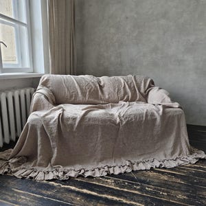 Peut inclure: Une housse de canap&eacute; en lin beige drap&eacute;e sur un canap&eacute;, pr&eacute;sentant une surface en tissu textur&eacute; et un ourlet &agrave; volants qui s'&eacute;tend le long du bord inf&eacute;rieur. La housse couvre enti&egrave;rement l'assise, le dossier et les accoudoirs avec un ajustement ample.