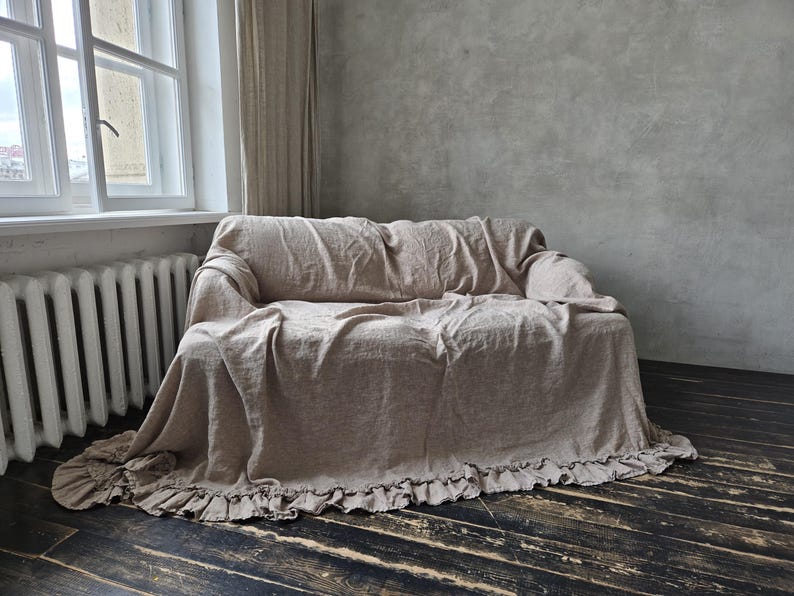 Peut inclure: Une housse de canap&eacute; en lin beige drap&eacute;e sur un canap&eacute;, pr&eacute;sentant une surface en tissu textur&eacute; et un ourlet &agrave; volants qui s'&eacute;tend le long du bord inf&eacute;rieur. La housse couvre enti&egrave;rement l'assise, le dossier et les accoudoirs avec un ajustement ample.