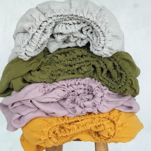 May include: A stack of four fitted bed sheets in various colors: light gray, olive green, lavender, and mustard yellow. The sheets are made of a soft, textured fabric and are neatly folded, showcasing their elasticized edges.