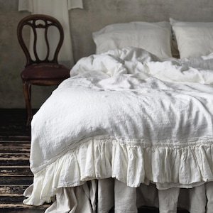 Könnte beinhalten: Ein Bett mit weißer Leinenbettwäsche, einschließlich eines gerüschten Bettrands und Kissen. Im Hintergrund steht ein Holzstuhl. Der Raum hat eine neutrale Farbpalette mit einer rustikalen, Vintage-Ästhetik.
