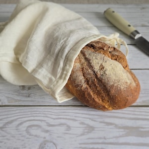 May include: A loaf of rustic bread, partially wrapped in a cream-colored linen cloth, rests on a light wood surface. A bread knife with a cream handle lies nearby. The bread has a golden-brown crust.