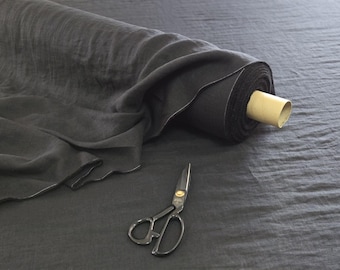 May include: A roll of dark gray fabric with a section unrolled, revealing the texture. A pair of scissors with gold accents lies nearby. The fabric appears to be linen or a similar material, suitable for sewing or crafting projects.