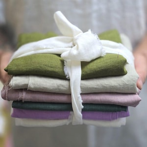 May include: A stack of colorful linen fabric squares in shades of green, beige, pink, dark green, and purple, tied with a white ribbon. The fabric is neatly folded and presented, suggesting a craft or textile product.
