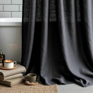 May include: Dark gray linen curtain hanging in front of a white bathtub. A stack of folded towels, a candle in a white mug, and a small brown mug sit on a woven mat. The scene evokes a spa-like atmosphere.