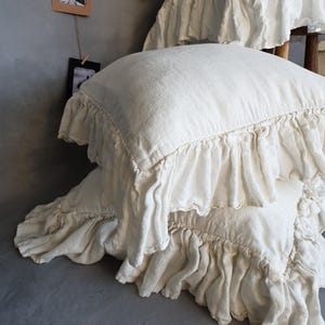 May include: Cream-colored linen pillows with ruffled edges. The pillows are stacked, showcasing the texture and design of the bedding. The ruffles add a decorative element to the otherwise simple design. The pillows are on a gray surface.