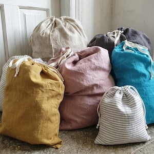 May include: A collection of linen drawstring bags in various colors, including mustard yellow, dusty rose, and teal. The bags have white drawstrings and are made of a textured fabric. Some bags have a striped pattern.