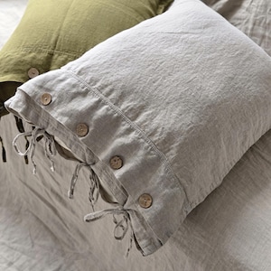 May include: Two linen pillowcases in a bedroom setting. One is light gray with wooden buttons and ties, the other is olive green. The pillowcases are on a bed with a neutral-colored duvet.