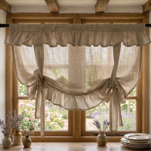 May include: A beige linen window curtain featuring a ruffled top valance and two tie-up sections that create a gathered, arched bottom edge. The curtain is hung on a black rod over a wooden window frame, allowing soft light to filter through the fabric.