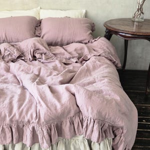 May include: A bed with a dusty rose linen duvet cover and matching ruffled pillowcases. The bed skirt is a light beige color. A dark wood side table with a vase of branches sits nearby.