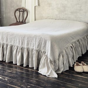 Puede incluir: Una falda de cama de lino gris claro con un borde con volantes. La falda de cama está sobre un suelo de madera.