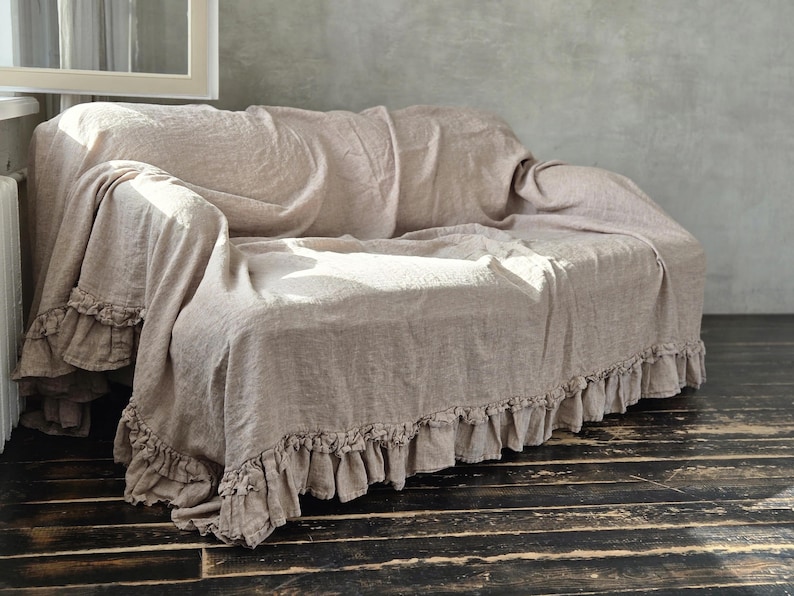 Peut inclure: Une housse de canap&eacute; beige avec une finition en tissu textur&eacute;, dot&eacute;e d'un bord &agrave; volants sur le bas. La housse recouvre enti&egrave;rement le canap&eacute;, y compris les accoudoirs et le dossier, avec une coupe ample et fluide cr&eacute;ant des plis souples.