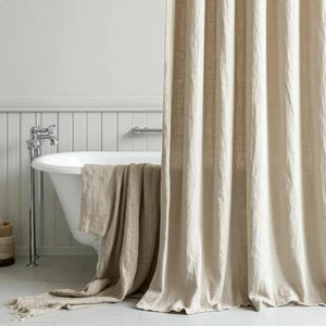 K&ouml;nnte beinhalten: Ein beigefarbener Duschvorhang aus Leinen mit sichtbarer Webstruktur h&auml;ngt vertikal und verdeckt teilweise eine wei&szlig;e Badewanne. Ein passendes beiges Leinenhandtuch mit ausgefransten Kanten liegt &uuml;ber dem Wannenrand und zeigt eine nat&uuml;rliche Textur.