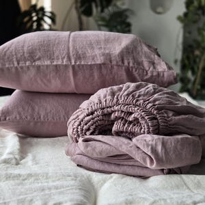 May include: A set of mauve-colored linen bedding, including two pillowcases and a fitted sheet, is neatly arranged on a white bed. The bedding has a soft, textured appearance, suggesting a comfortable and relaxed aesthetic. The scene is illuminated by natural light.