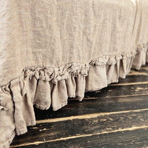 Peut inclure: Un cache-sommier en lin beige dot&eacute; d'une bordure &agrave; double volant sur le bas. Le tissu pr&eacute;sente une texture tiss&eacute;e visible et une teinte naturelle, tombant vers le bas pour cr&eacute;er un aspect fronc&eacute; et doux le long de la base.