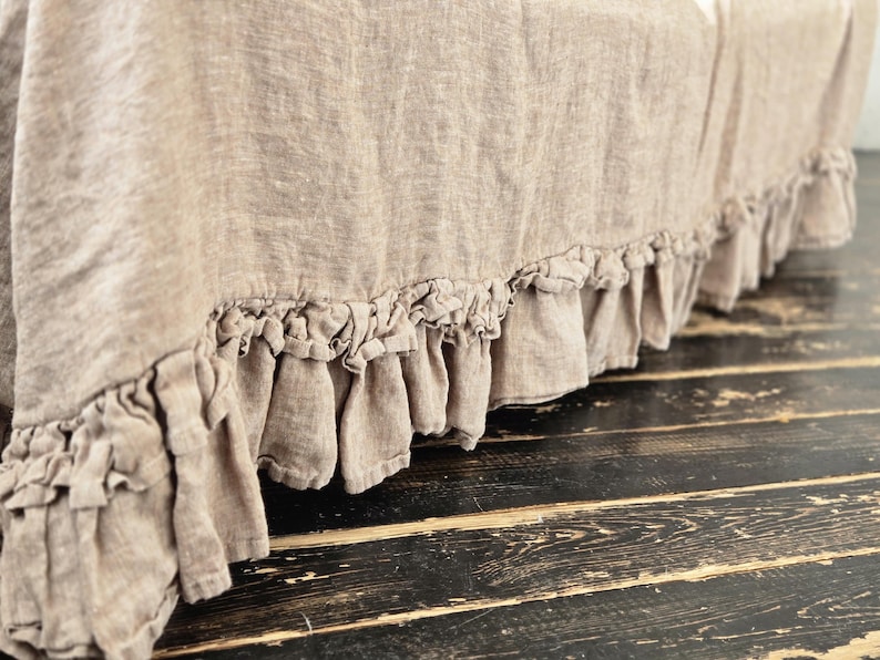 Peut inclure: Un cache-sommier en lin beige dot&eacute; d'une bordure &agrave; double volant sur le bas. Le tissu pr&eacute;sente une texture tiss&eacute;e visible et une teinte naturelle, tombant vers le bas pour cr&eacute;er un aspect fronc&eacute; et doux le long de la base.