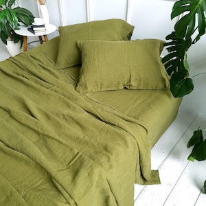 Puede incluir: Juego de cama de lino verde oliva, que incluye una sábana bajera y dos fundas de almohada. Las sábanas están drapeadas sobre una cama, con un aspecto texturizado. El color es un tono apagado y terroso, creando una estética relajante. La ropa de cama está rodeada de plantas.
