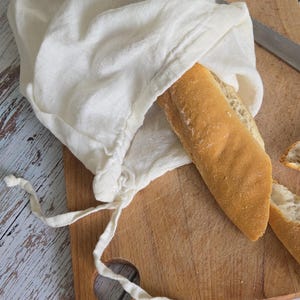 May include: A fresh baguette partially emerges from a white linen bread bag on a wooden cutting board. The bread has a golden-brown crust and a soft interior. A knife rests on the board, suggesting preparation for a meal.